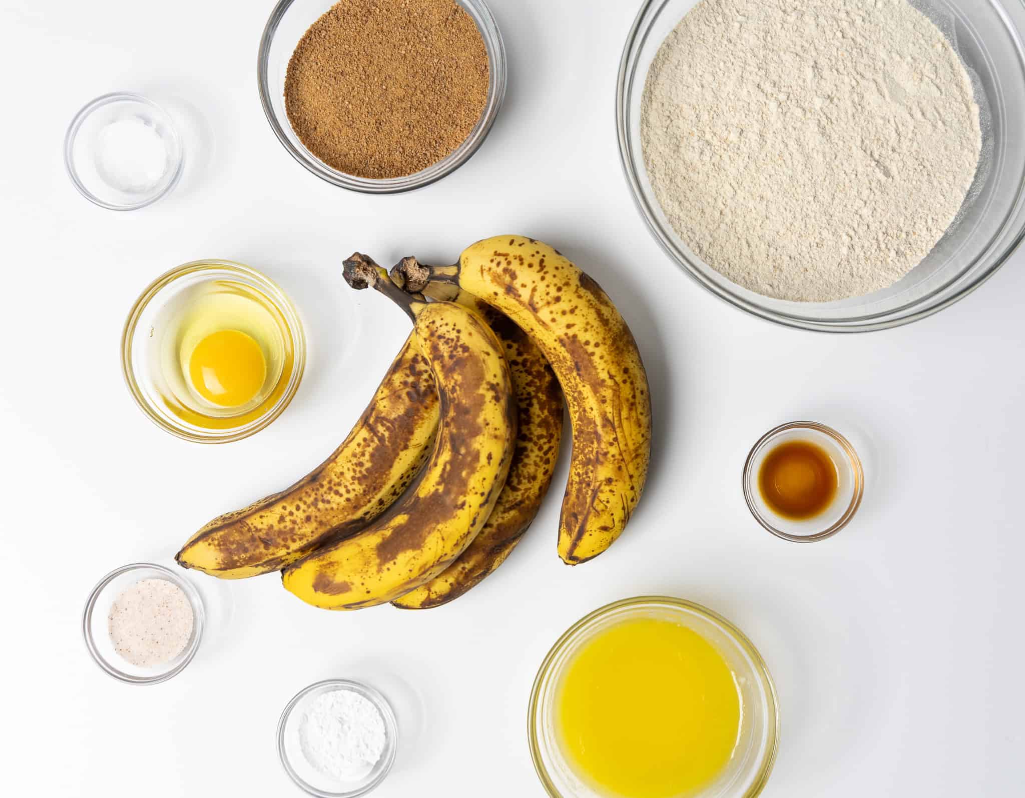 A collection of ingredients arranged on a white background, for making einkorn banana bread. At the center, there is a bunch of ripe bananas with brown spots. Surrounding them are various glass bowls containing different ingredients: A bowl of flour, a bowl of coconut sugar, a cracked egg, melted butter, vanilla extract, salt, baking powder, and baking soda.
