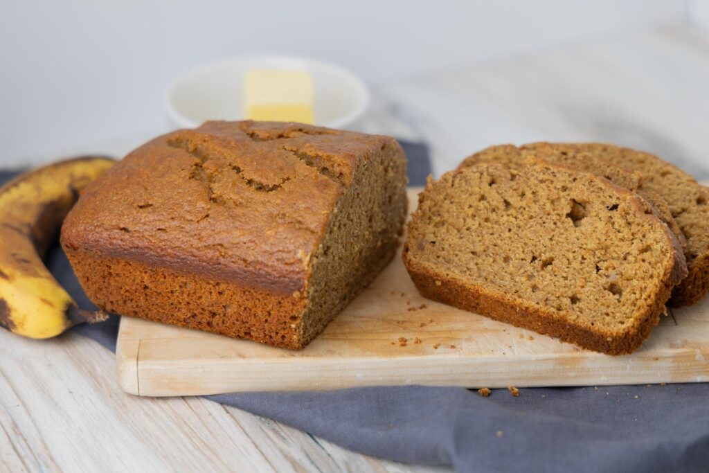 The image shows a freshly baked loaf of einkorn banana bread with a golden-brown crust, partially sliced to reveal its moist, textured interior. The bread is placed on a wooden cutting board, with a couple of slices arranged beside it. In the background, there is a ripe banana and a small dish containing a pat of butter. The setting has a cloth napkin adding to the warm and inviting presentation.