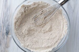 The image shows a clear glass bowl filled with a light brown, slightly coarse dry mixture. A stainless steel whisk is partially submerged in the mixture, indicating it is being combined. These dry ingredients are for making einkorn banana bread and include einkorn flour, baking soda, baking powder, salt, and coconut sugar.