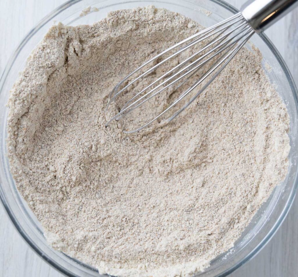 The image shows a clear glass bowl filled with a light brown, slightly coarse dry mixture. A stainless steel whisk is partially submerged in the mixture, indicating it is being combined. These dry ingredients are for making einkorn banana bread and include einkorn flour, baking soda, baking powder, salt, and coconut sugar. 