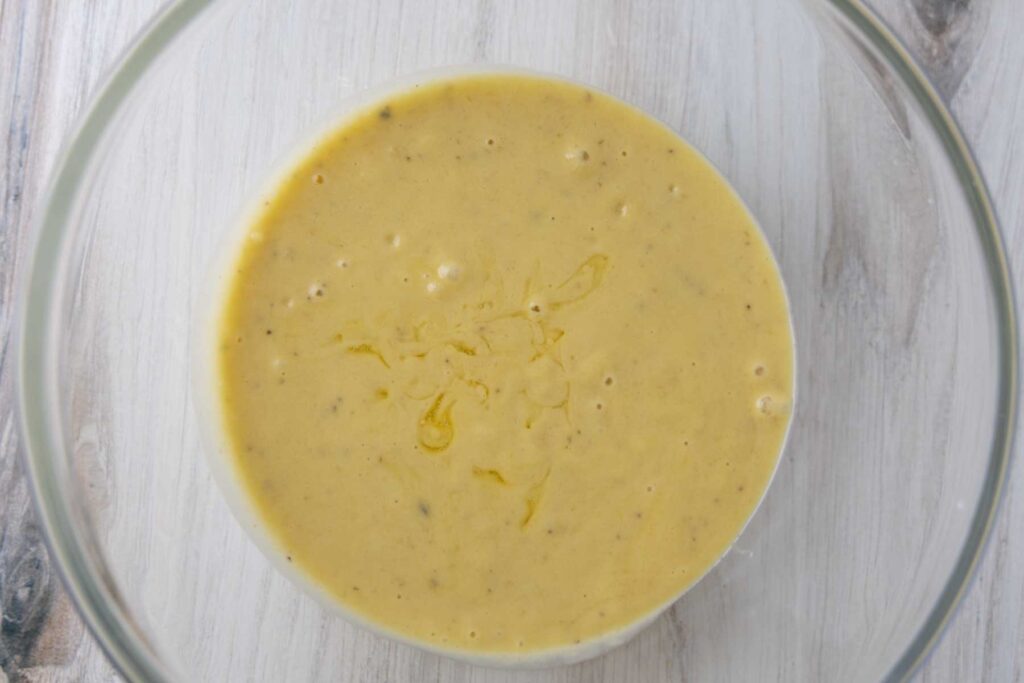 The image shows a clear glass bowl filled with a smooth, golden-yellow liquid mixture with small bubbles and specks throughout. This mixture consists of the wet ingredients for making einkorn banana bread, including an egg, melted butter, and mashed bananas. 
