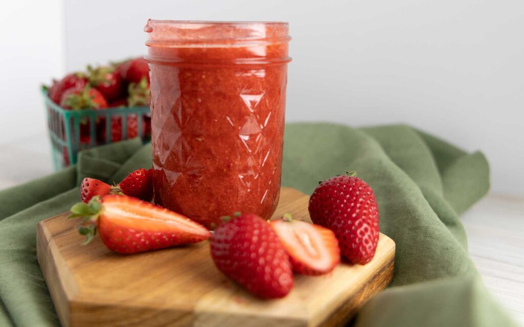 There's a jar of homemade strawberry jam placed prominently in the center, filled to the top with a vibrant, rich red spread that looks thick and freshly made. The jar has a textured diamond pattern and is sitting on a small wooden board, which rests atop a soft green cloth, adding a rustic, cozy touch. Surrounding the jar are fresh strawberries—some whole, others sliced open to reveal their juicy, red interiors. In the background, a green berry basket filled with more strawberries is slightly out of focus, reinforcing the fresh, farm-to-table vibe. The overall aesthetic is warm and inviting, perfect for showcasing homemade goodness.