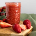 there's a jar of homemade strawberry jam placed prominently in the center, filled to the top with a vibrant, rich red spread that looks thick and freshly made. The jar has a textured diamond pattern and is sitting on a small wooden board, which rests atop a soft green cloth, adding a rustic, cozy touch. Surrounding the jar are fresh strawberries—some whole, others sliced open to reveal their juicy, red interiors. In the background, a green berry basket filled with more strawberries is slightly out of focus, reinforcing the fresh, farm-to-table vibe. The overall aesthetic is warm and inviting, perfect for showcasing homemade goodness.