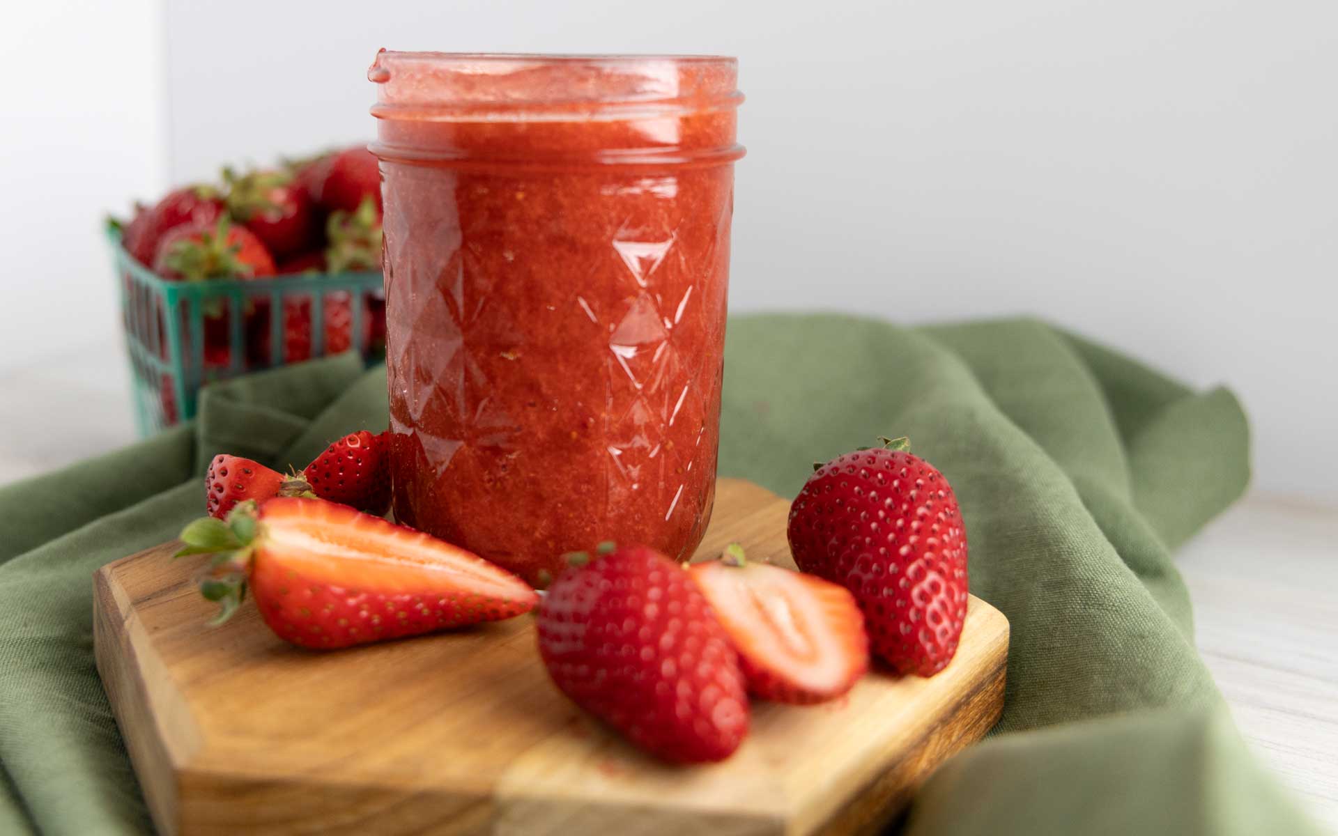 there's a jar of homemade strawberry jam placed prominently in the center, filled to the top with a vibrant, rich red spread that looks thick and freshly made. The jar has a textured diamond pattern and is sitting on a small wooden board, which rests atop a soft green cloth, adding a rustic, cozy touch. Surrounding the jar are fresh strawberries—some whole, others sliced open to reveal their juicy, red interiors. In the background, a green berry basket filled with more strawberries is slightly out of focus, reinforcing the fresh, farm-to-table vibe. The overall aesthetic is warm and inviting, perfect for showcasing homemade goodness.