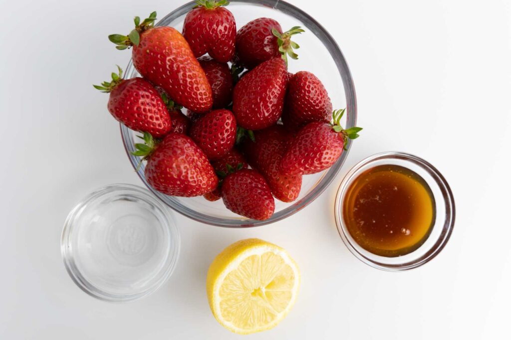 In this image, we see the simple, wholesome ingredients used to make homemade strawberry jam. A clear glass bowl is filled with fresh, ripe strawberries, their vibrant red color and glossy skin making them the star of the scene. Beside them sits a small bowl of golden honey, thick and rich, ready to add natural sweetness. A halved lemon, juicy and bright, hints at the fresh citrus flavor that will balance the sweetness and help the jam set. Finally, a small glass bowl of water —completes the lineup. The clean white background and natural lighting highlight the freshness and simplicity of each ingredient, perfect for a nourishing, homemade preserve.