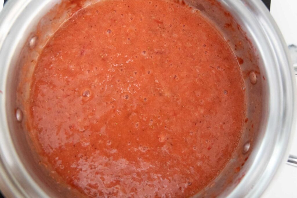This image shows a pot filled with homemade strawberry jam. The mixture has a thick, slightly chunky texture and a rich reddish-pink color, with small bubbles and bits of fruit visible throughout. It is in the cooking stage.