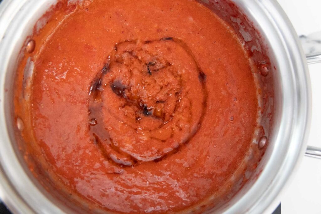 This image shows a pot of homemade strawberry jam in the process of being sweetened or flavored. The jam has a thick, smooth consistency and a rich reddish color, similar to the previous photo, but now there’s a swirl of honey and lemon juice being stirred in. The swirl pattern shows the ingredients haven’t fully mixed in yet, giving a visual cue that the jam is still being prepared.