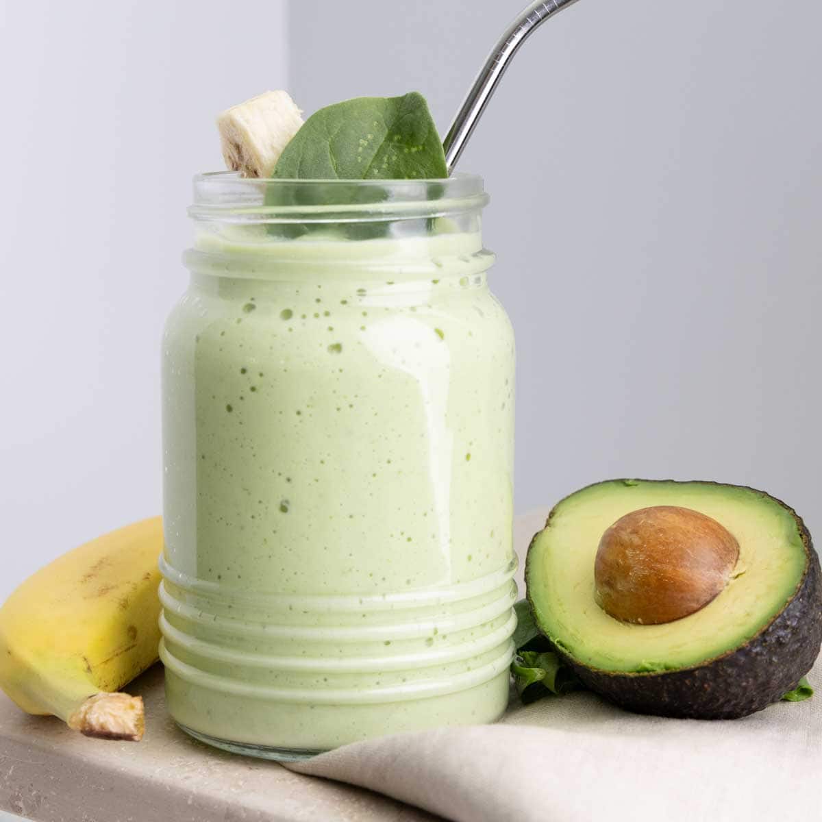 A creamy green avocado banana smoothie served in a glass jar with a metal straw. The smoothie is topped with a fresh spinach leaf and a small banana slice. In the background, a whole banana and a halved avocado with the pit are placed on a light surface, with a neutral-colored napkin nearby. The image has a clean and minimal aesthetic, highlighting the fresh, wholesome ingredients used to make this nourishing smoothie.