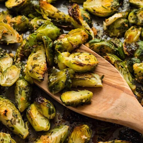 Golden brown oven roasted Brussels sprouts with garlic and herbs, served on a baking sheet with a wooden spoon.