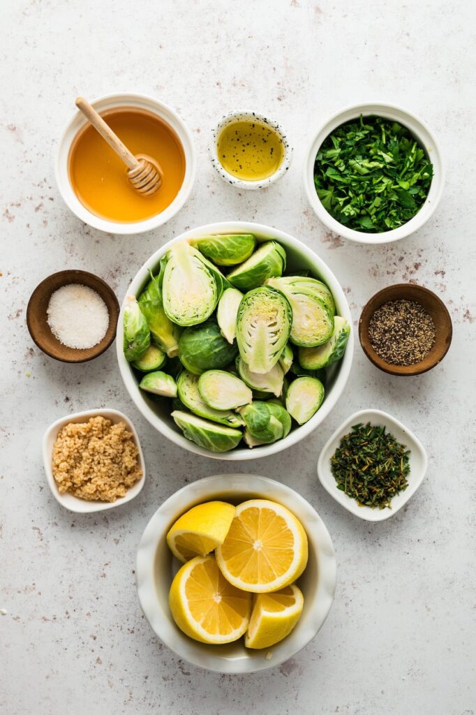 Fresh Brussels sprouts, lemon, garlic, parsley, thyme, honey, olive oil, salt, and pepper arranged in bowls on a white surface for oven roasted Brussels sprouts.