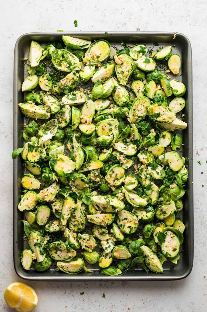 Baking sheet filled with halved Brussels sprouts tossed with garlic, parsley, thyme, salt, pepper, olive oil, and honey, ready to roast in the oven