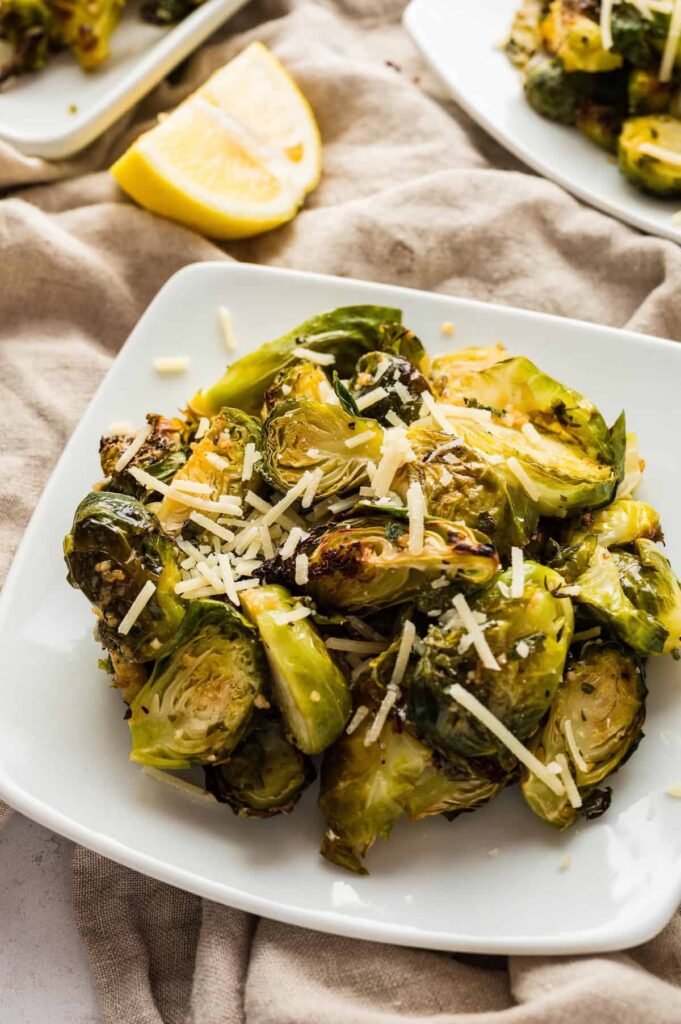 Oven roasted Brussels Sprouts served on a white plate, topped with freshly grated Parmesan cheese and paired with lemon wedges.