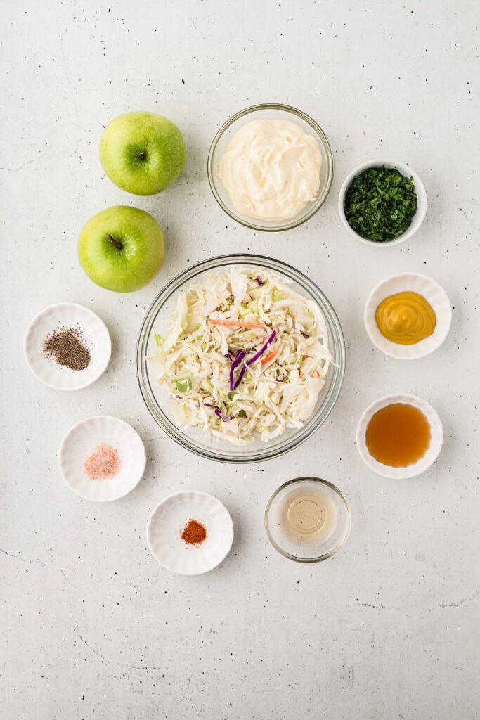 Overhead view of apple coleslaw ingredients including green apples, coleslaw mix, mayonnaise, chopped cilantro, mustard, honey, apple cider vinegar, salt, pepper, and cayenne pepper in small bowls on a white background.”