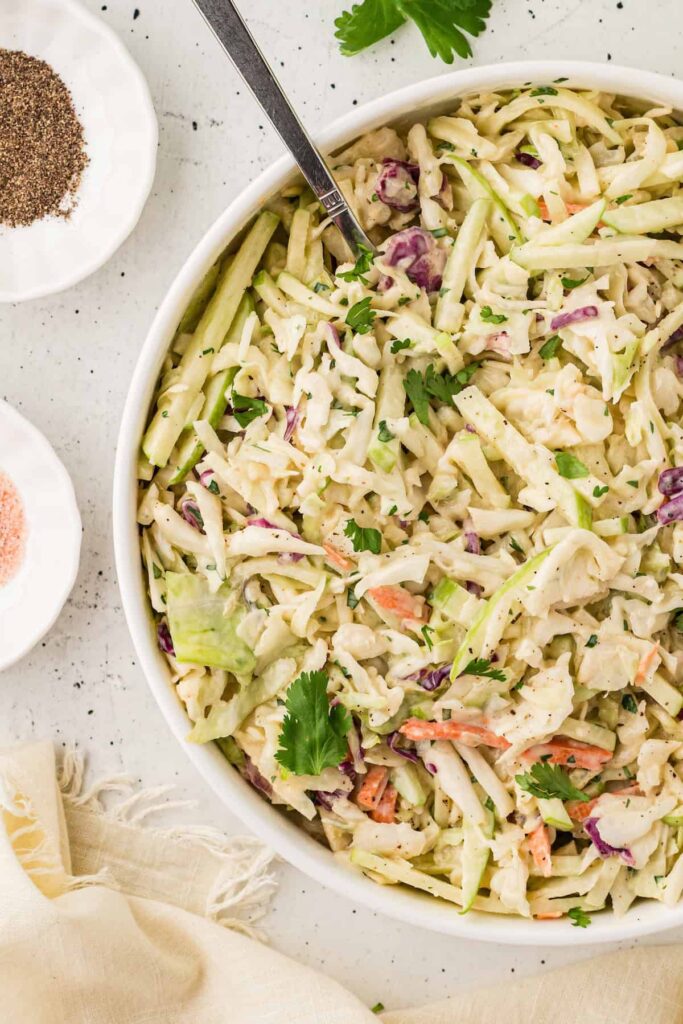 A large white bowl filled with creamy apple coleslaw made of shredded cabbage, green apple matchsticks, carrots, and fresh cilantro. The slaw is tossed in a honey-mustard dressing, with sprigs of cilantro on top. Small dishes of black pepper and pink salt sit beside the bowl on a white surface with a beige napkin.