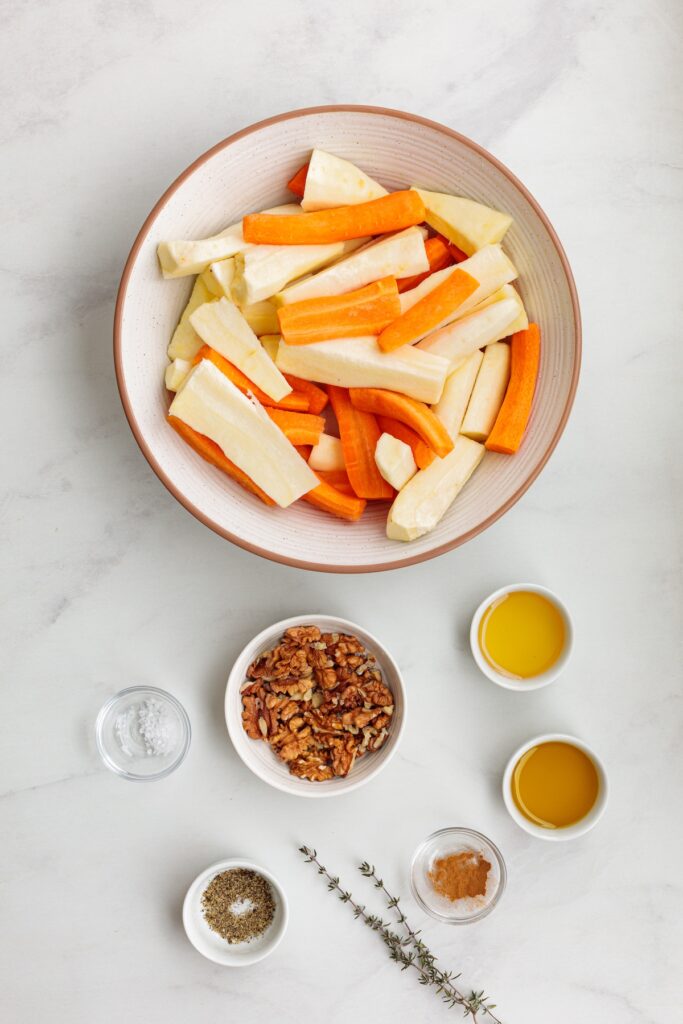 Overhead view of recipe ingredients for roasted honey carrots and turnips, including a large bowl of peeled and cut carrots and turnips, small bowls of walnuts, avocado  oil, honey, salt, pepper, cinnamon, and sprigs of fresh thyme on a white surface.