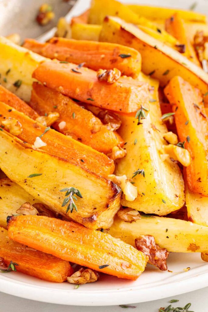 Close-up of roasted honey-glazed carrots and turnips on a white plate, garnished with fresh thyme and chopped walnuts. The vegetables are golden, caramelized, and slightly crisp around the edges