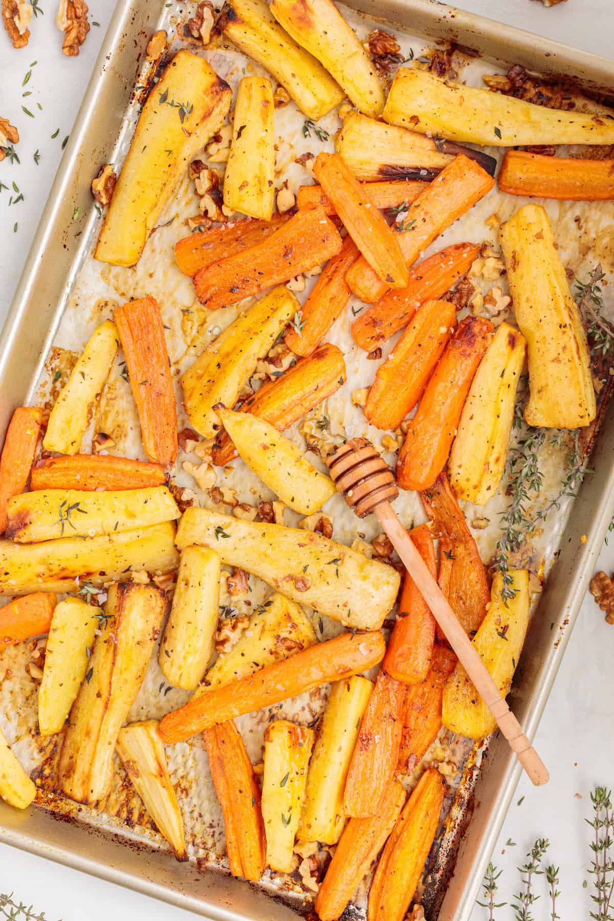 Golden roasted carrot and parsnip sticks on a parchment-lined baking sheet, sprinkled with toasted walnuts and fresh thyme, with a honey dipper resting on top for a touch of sweetness.