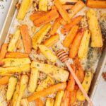 Golden roasted carrot and parsnip sticks on a parchment-lined baking sheet, sprinkled with toasted walnuts and fresh thyme, with a honey dipper resting on top for a touch of sweetness.
