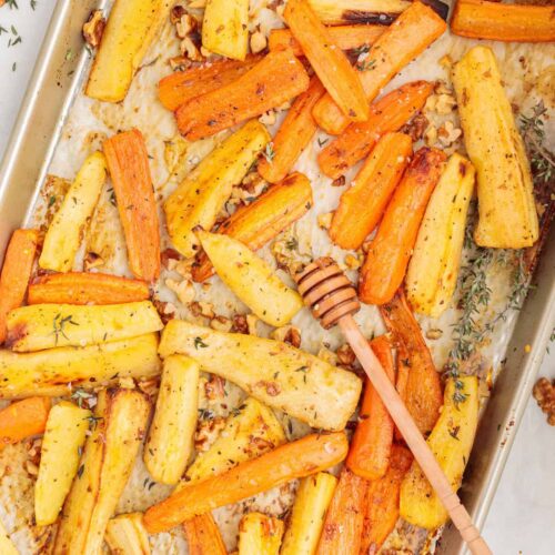 Golden roasted carrot and parsnip sticks on a parchment-lined baking sheet, sprinkled with toasted walnuts and fresh thyme, with a honey dipper resting on top for a touch of sweetness.