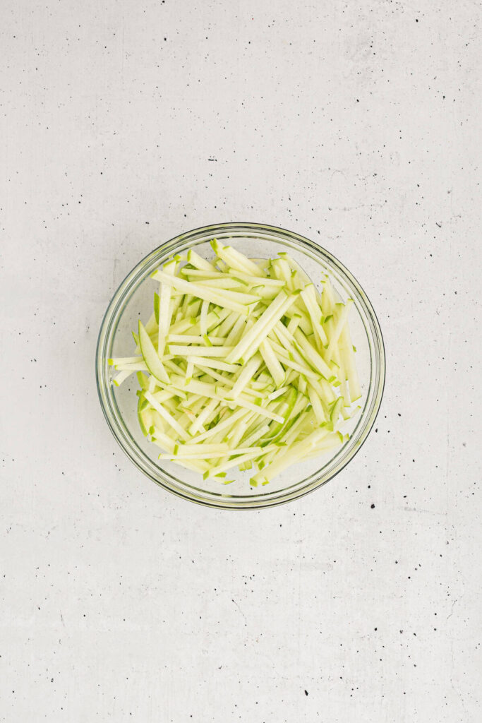 Glass bowl filled with thin matchstick slices of fresh green apples, prepped for making apple coleslaw.
