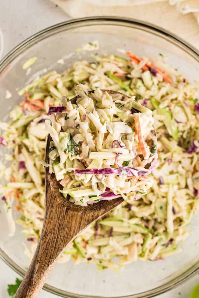 Wooden spoon scooping up creamy apple coleslaw from a glass mixing bowl, showing shredded cabbage, carrots, green apples, and herbs