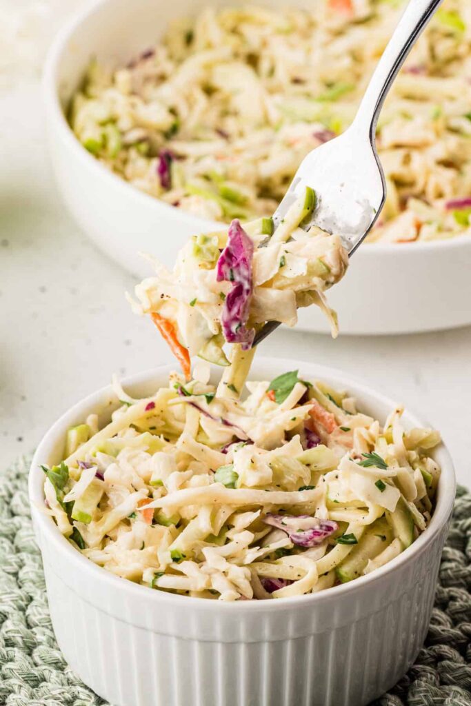 Close-up of creamy apple coleslaw in a white ramekin, with a fork lifting a bite showing cabbage, carrots, green apples, and cilantro.