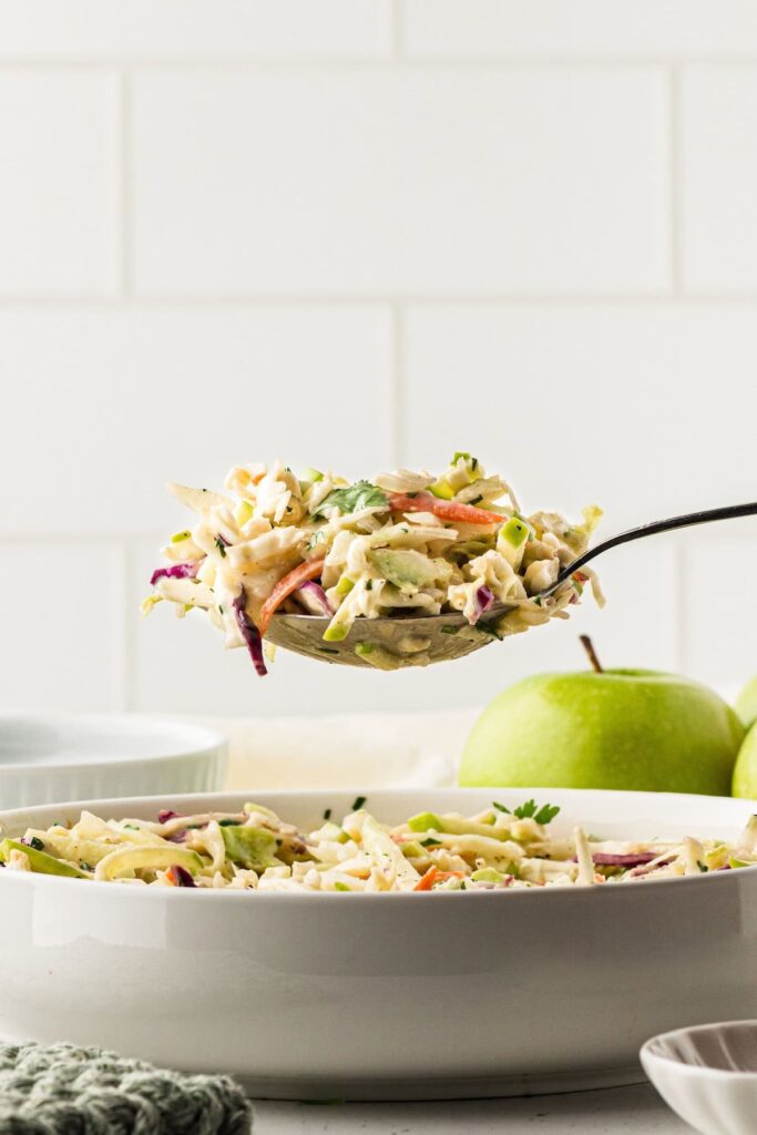 Spoon lifting a serving of creamy apple coleslaw with cabbage, carrots, and fresh herbs from a white bowl, with green apples in the background.