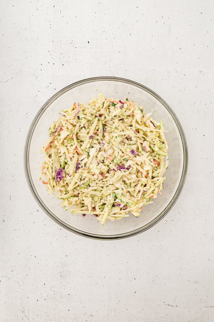 A clear glass bowl filled with apple coleslaw made of shredded cabbage, carrots, green apple matchsticks, and purple cabbage, all tossed in a creamy dressing. The bowl sits on a light speckled surface.
