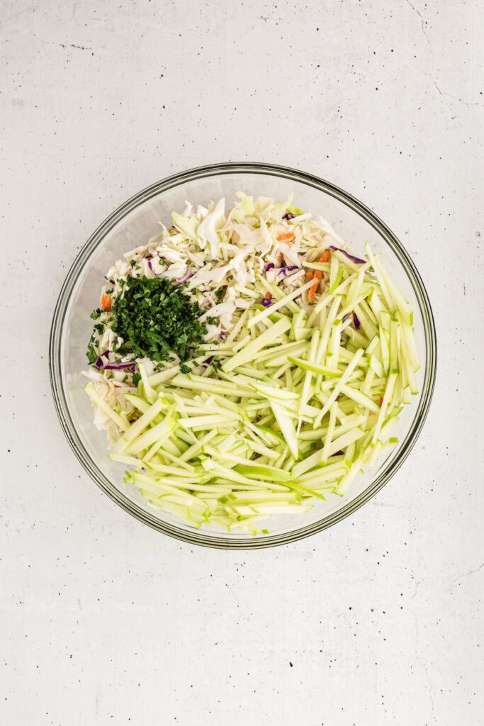 Large glass bowl with shredded cabbage, matchstick green apples, and chopped cilantro ready to be mixed for homemade apple coleslaw.
