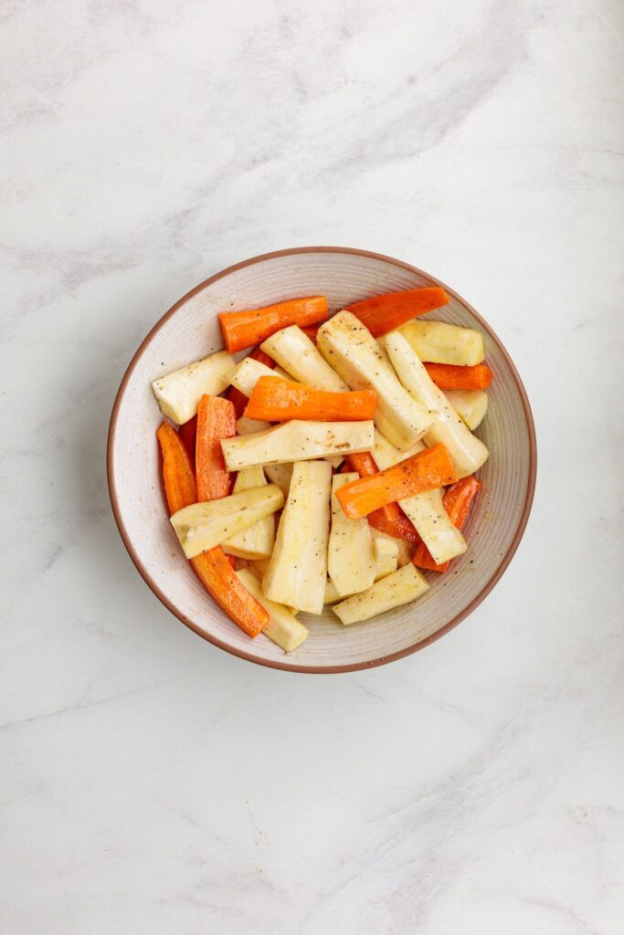 Overhead view of a bowl filled with peeled and chopped carrots and turnips, lightly seasoned and ready to be roasted.