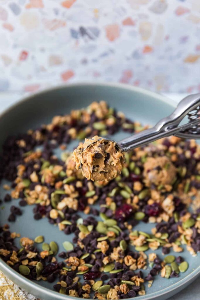A metal cookie scoop holds a portion of peanut butter energy ball mixture with chocolate chips, nuts, and dried fruit. The scoop hovers over a blue plate scattered with granola, pumpkin seeds, dried cranberries, and mini chocolate chips.