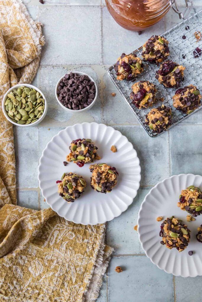 Homemade peanut butter energy balls rolled with chocolate chips, pepitas, granola, and dried cranberries, served on white plates and a cooling rack. Small bowls of chocolate chips and peptias, a jar of honey, and a mustard-yellow patterned cloth are arranged around the setup on a tiled surface.