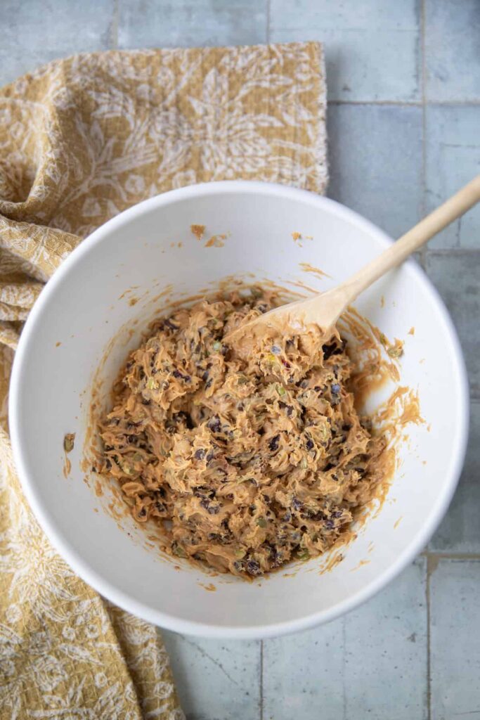 A white mixing bowl filled with a thick peanut butter mixture blended with dried cranberries, pistachios, seeds, and chocolate chips. A wooden spoon rests in the bowl, and a mustard-yellow patterned cloth is draped beside it on a tiled surface.