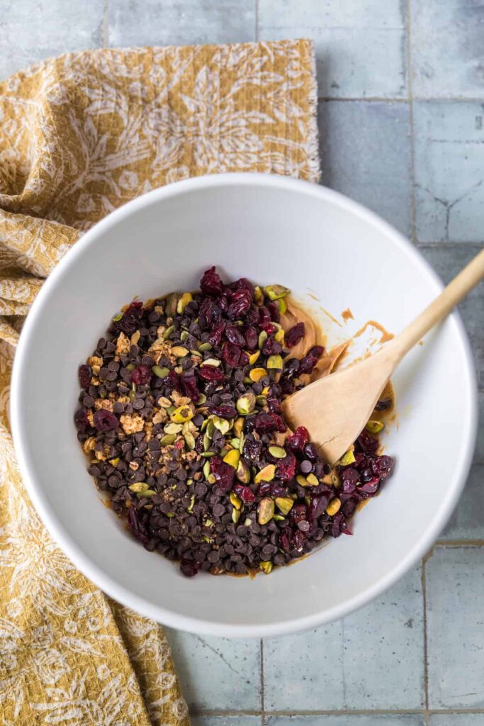 A white mixing bowl filled with peanut butter, chocolate chips, dried cranberries, pistachios, and seeds being stirred with a wooden spoon. A mustard-yellow patterned cloth sits beside the bowl on a tiled surface.