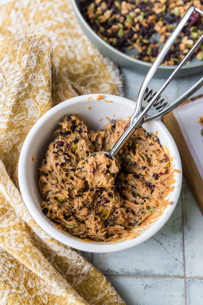 A white bowl filled with peanut butter mixture for protein balls, mixed with chocolate chips, dried cranberries, and pistachios. A metal cookie scoop rests in the mixture, ready for portioning. A mustard-yellow patterned cloth and tray with parchment paper sit nearby.