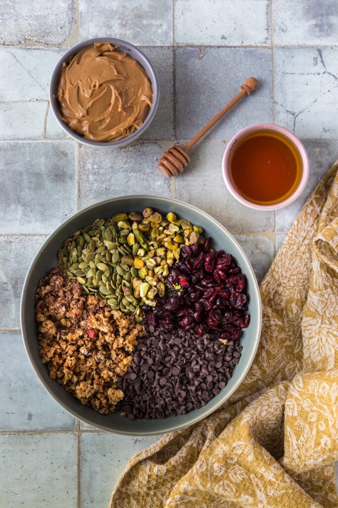 Overhead view of ingredients for peanut butter energy balls, including a bowl of pumpkin seeds (pepitas), pistachios, dried cranberries, granola, and mini chocolate chips. Nearby are small bowls of creamy peanut butter and honey with a wooden honey dipper, set on a tiled surface with a mustard-yellow patterned cloth.
