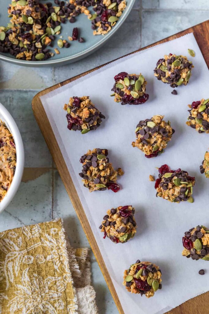 Overhead view of homemade no-bake peanut butter balls rolled in granola, pepitas, dried cranberries, and mini chocolate chips, arranged neatly on a parchment-lined wooden board with extra coating ingredients in a dish nearby.