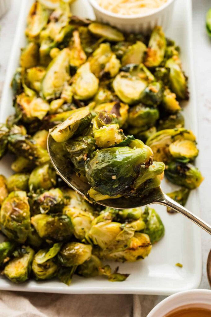 Close-up of oven-roasted Brussels sprouts served on a white platter, with a spoon lifting a portion to show their golden-brown, crispy edges.