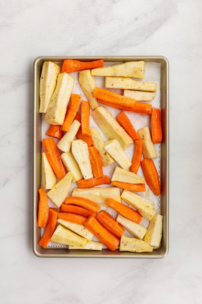 Chopped carrots and turnips spread out in a single layer on a parchment-lined baking sheet, seasoned and ready to be roasted