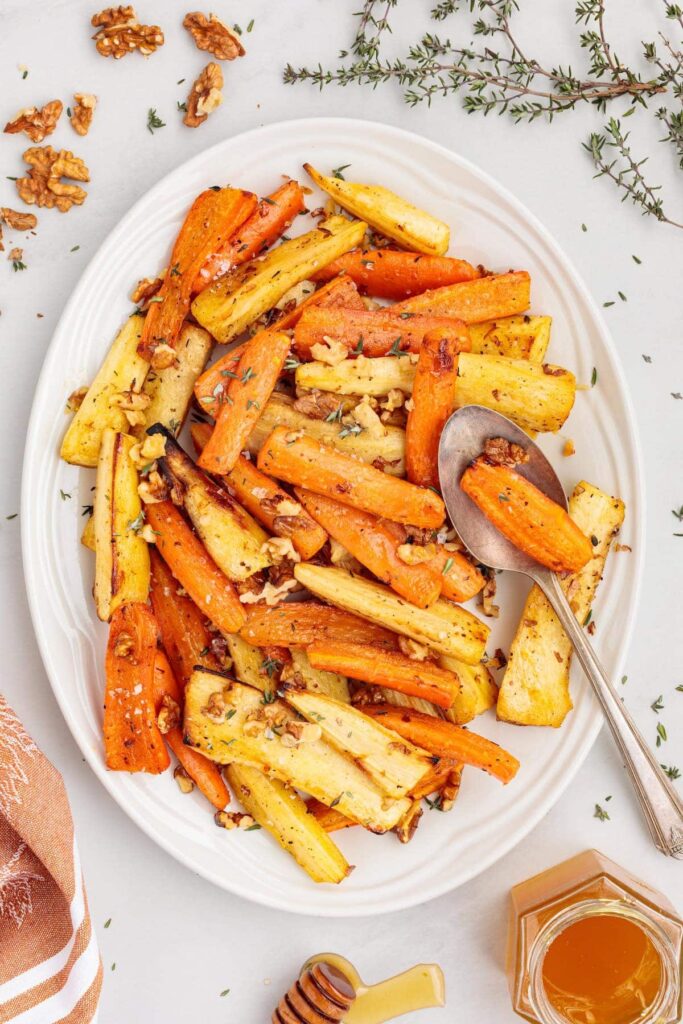 White serving platter filled with roasted carrots and parsnips tossed with walnuts, fresh thyme, and honey, with a spoon resting on the side.