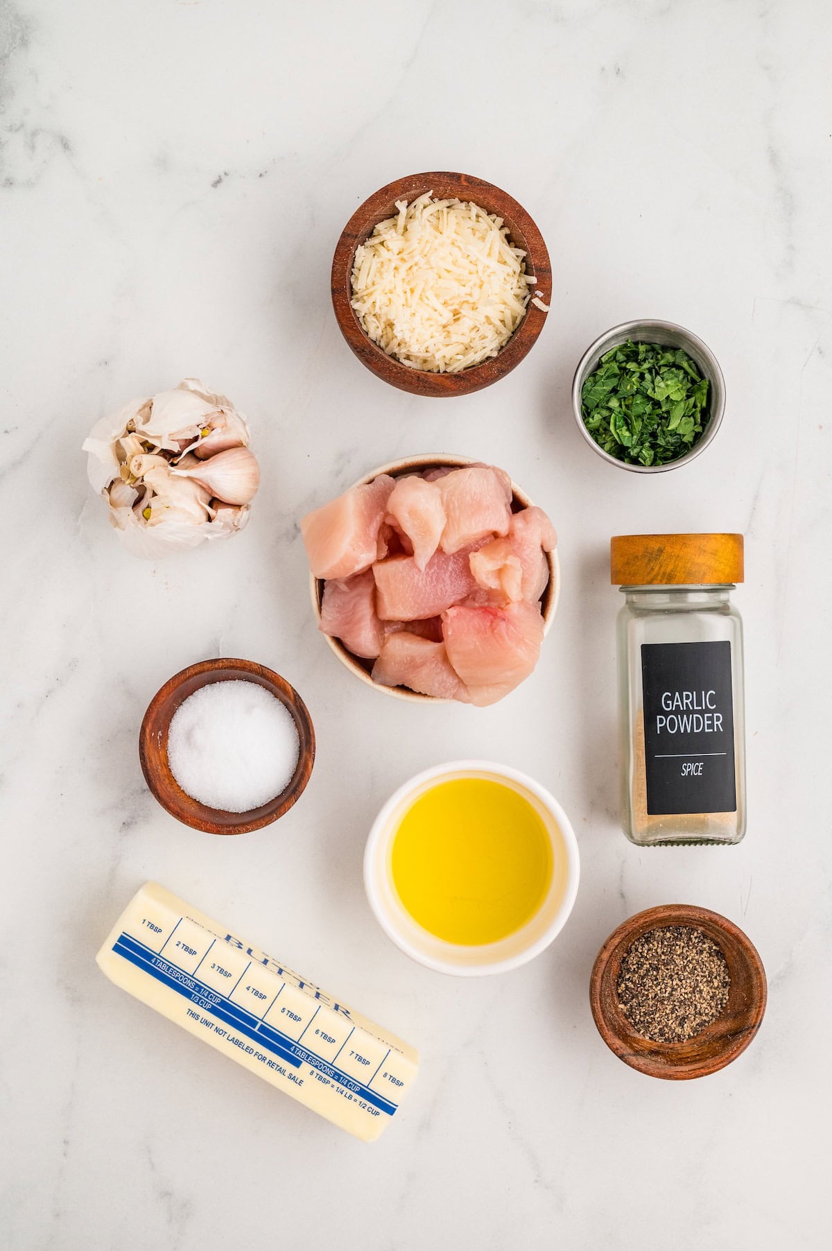 Top-down view of ingredients for One-Pan Garlic Parmesan Chicken Bites arranged on a white marble surface. Includes raw chicken pieces, grated parmesan cheese, chopped parsley, garlic bulb, garlic powder, salt, pepper, butter, and a small bowl of avocado oil — all neatly displayed in small bowls and dishes.