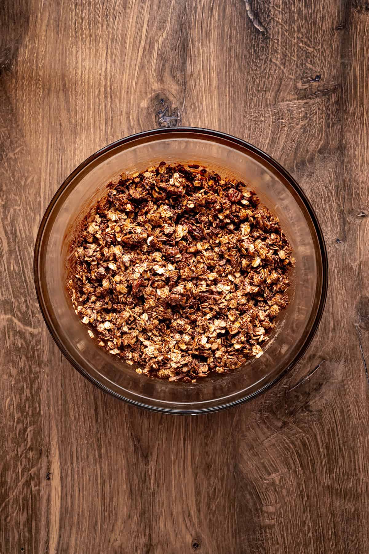 Glass bowl filled with mixed chocolate coconut granola ingredients coated in maple syrup vanilla extract, and coconut oil, ready to bake.