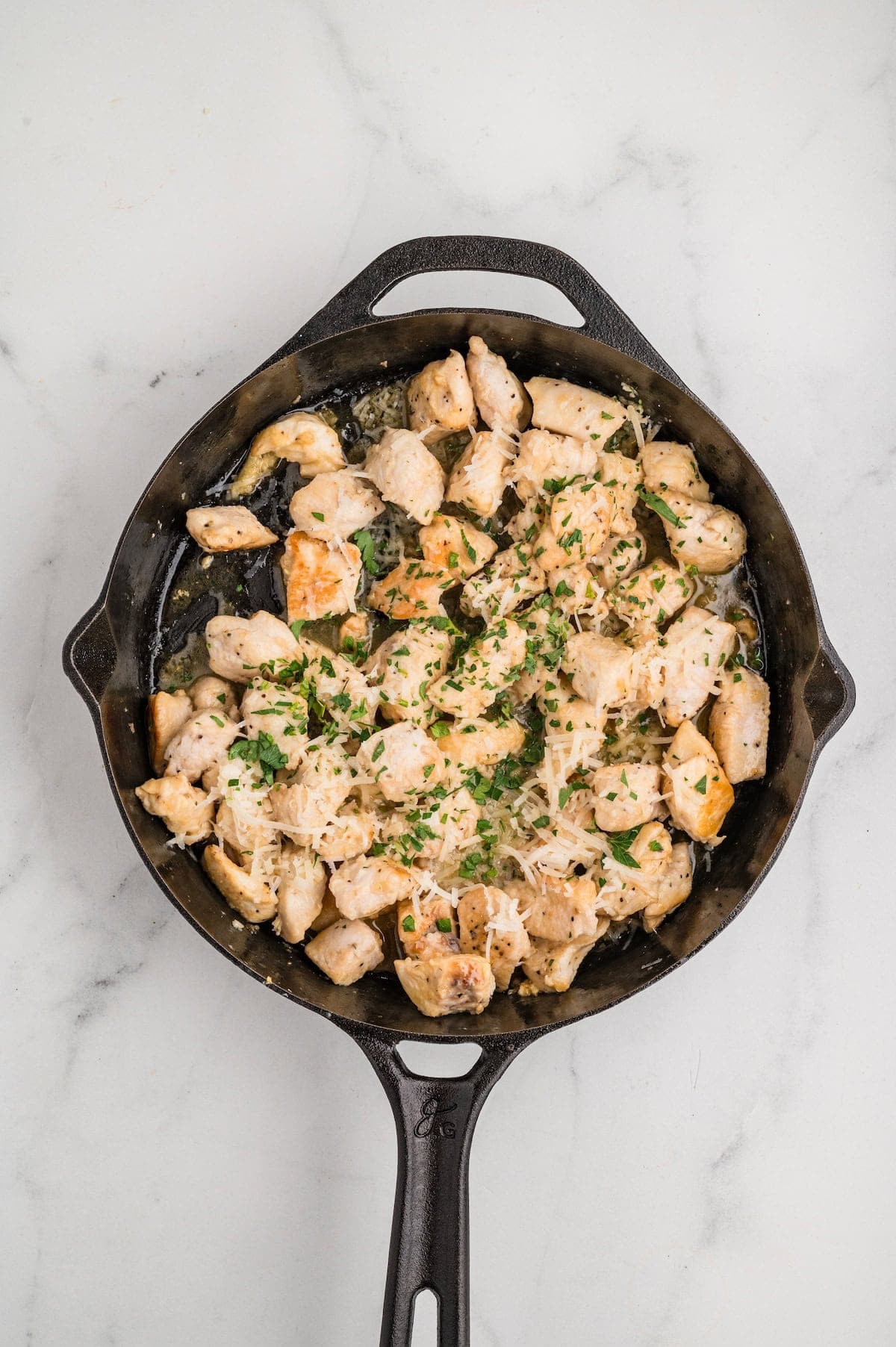Top-down view of finished Garlic Parmesan Chicken Bites in a cast iron skillet. The chicken pieces are golden and coated in a buttery garlic sauce, topped with freshly grated parmesan cheese and chopped parsley for garnish.