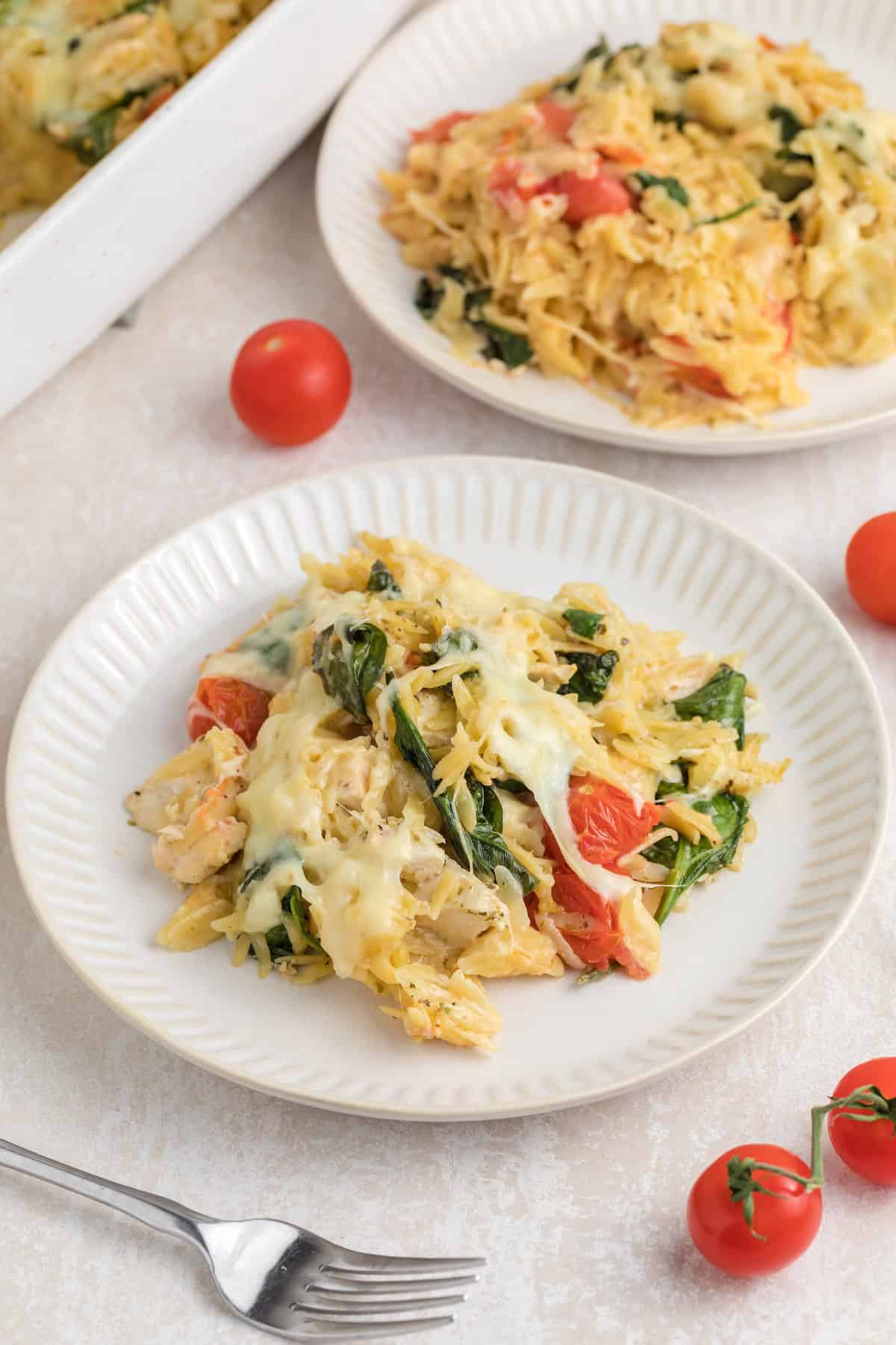 A serving of creamy baked chicken orzo with spinach, cherry tomatoes, and melted cheese on a white plate, with extra portions and fresh tomatoes in the background.