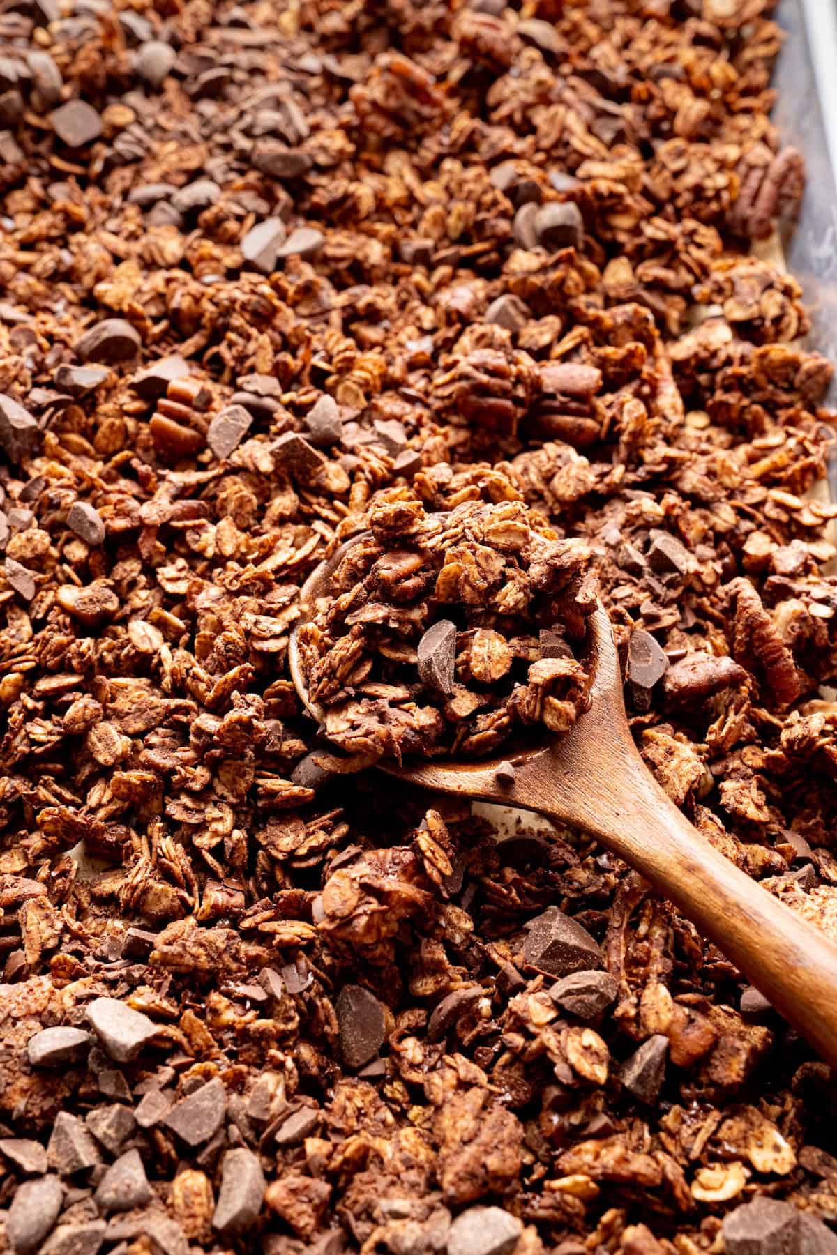 Wooden spoon scooping homemade chocolate coconut granola with oats, pecans, and dark chocolate chunks from a baking sheet.