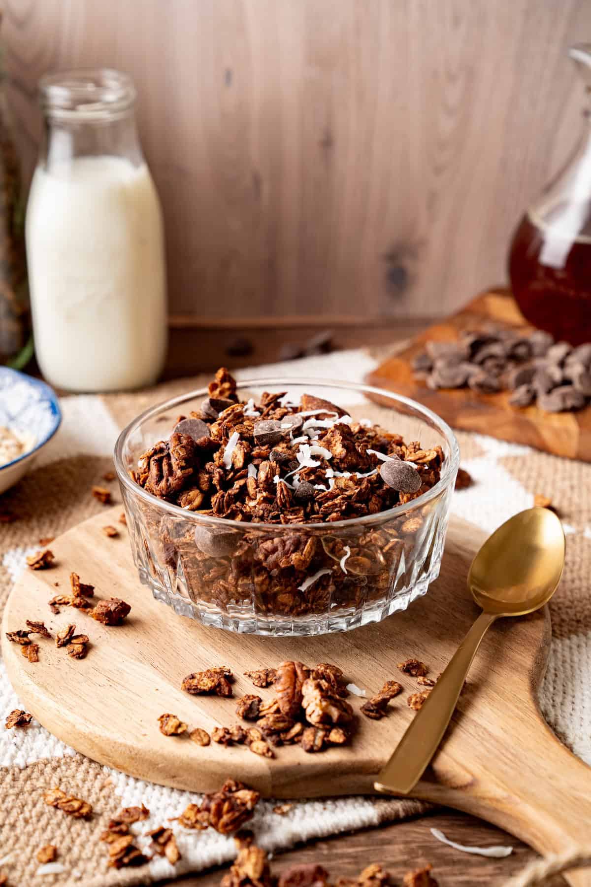 Glass bowl of homemade chocolate coconut granola topped with dark chocolate chunks and shredded coconut, served on a wooden board with a gold spoon, with milk and ingredients in the background.