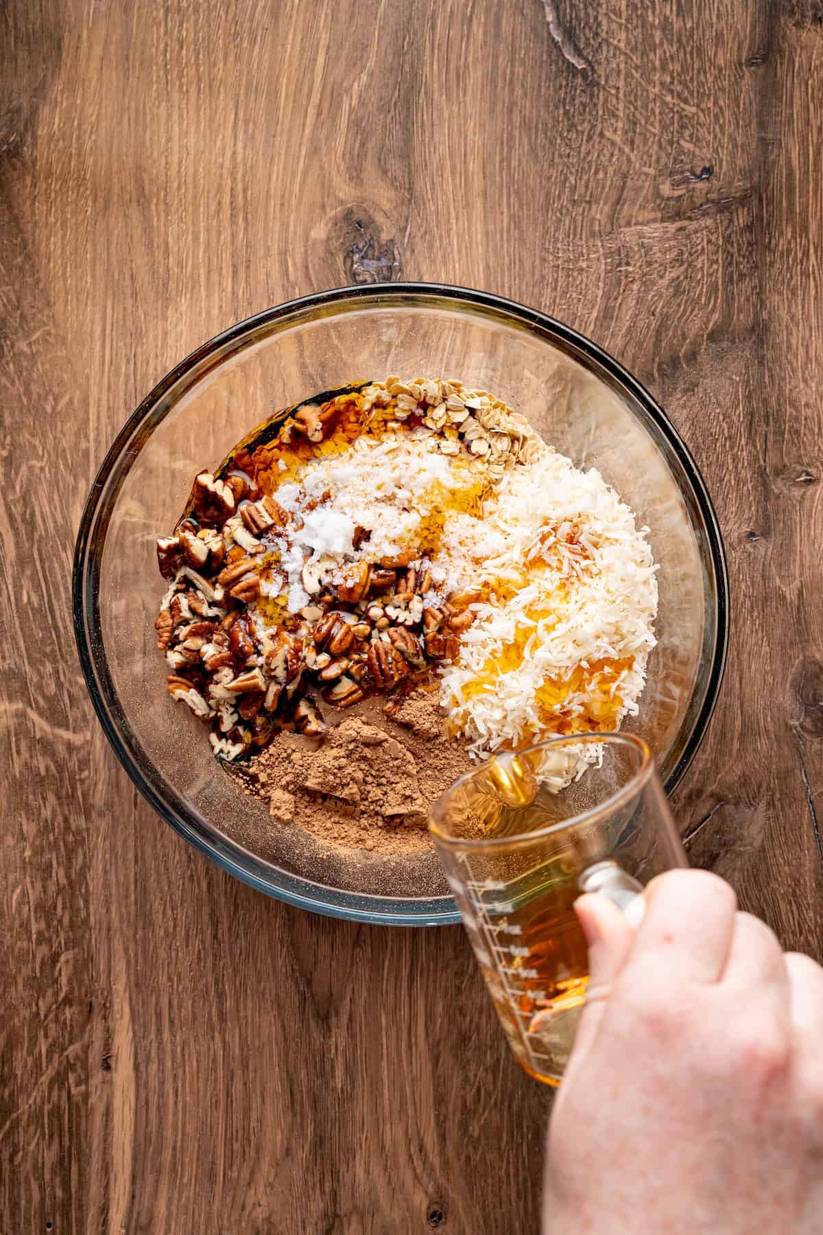 Hand pouring maple syrup into a glass mixing bowl with oats, shredded coconut, chopped pecans, cocoa powder, coconut oil, and vanilla extract to make homemade chocolate coconut granola.