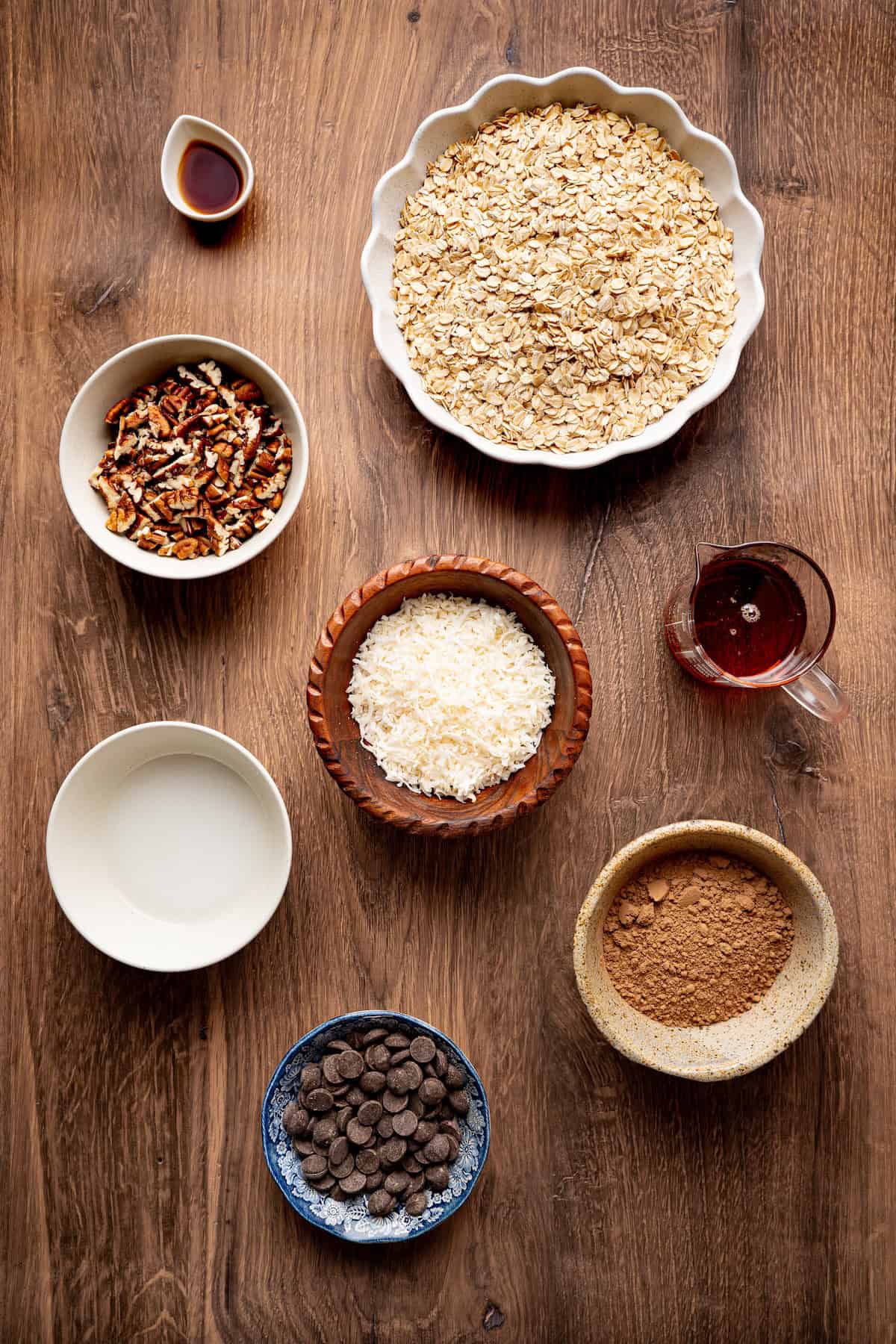 Overhead view of ingredients for homemade chocolate coconut granola, including rolled oats, cocoa powder, shredded coconut, chopped pecans, dark chocolate chips, maple syrup, coconut oil, and vanilla extract arranged in bowls on a wooden table.