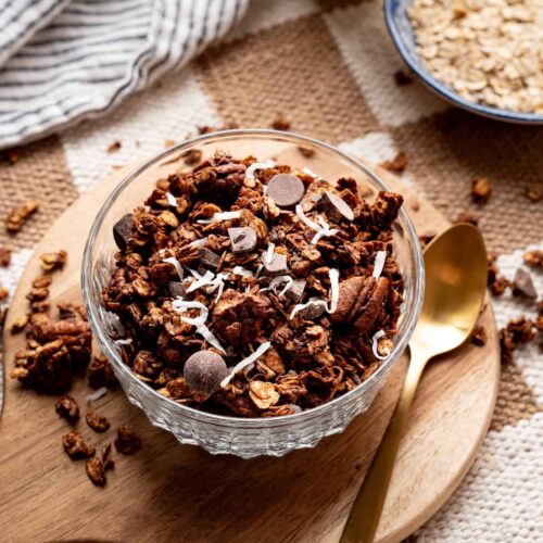 Glass bowl filled with homemade chocolate coconut granola topped with dark chocolate chips and shredded coconut, served on a wooden board with a gold spoon.