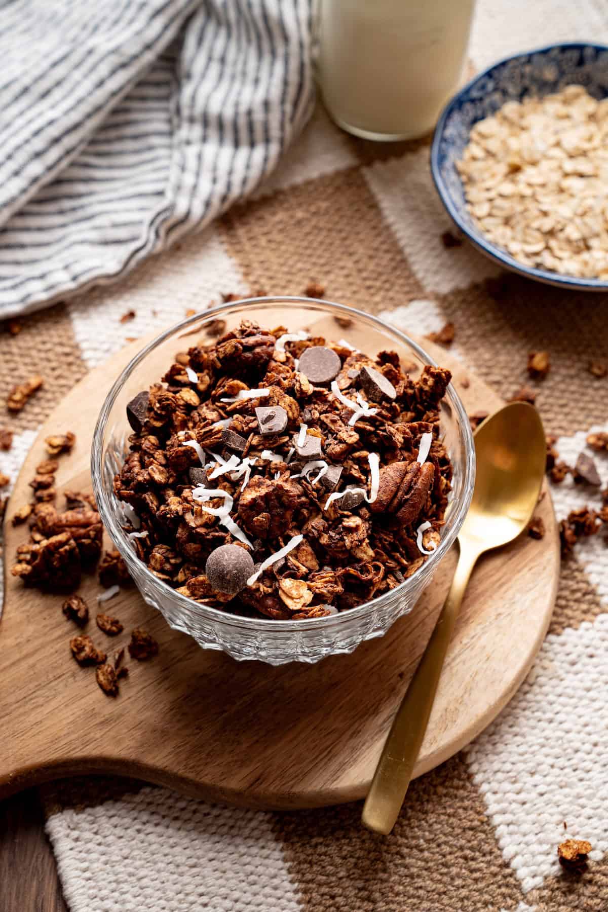 Glass bowl filled with homemade chocolate coconut granola topped with dark chocolate chips and shredded coconut, served on a wooden board with a gold spoon.